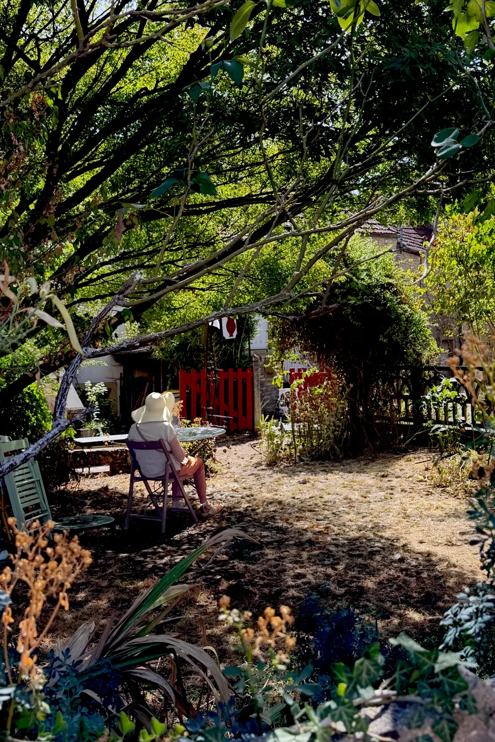 A woman wearing a wide-brimmed straw hat is sitting in a garden chair under a tree in the shade. The viewer sees her from behind